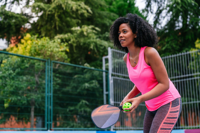 Learn what the perfect pickleball outfit looks like. A woman of color holds a pickleball paddle and ball, ready to hit the ball. She wears a pink athletic outfit to play pickleball. 