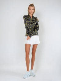 Woman wearing fitted camo print half-zip performance top with gold collar accents, thumbhole sleeves, and silver zipper detail, showcasing the versatile high collar design suitable for athletic and casual wear - front view with skirt