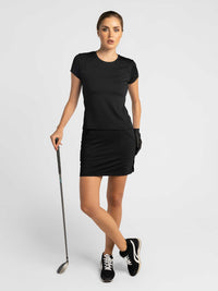 Front view of model wearing the womens golf 15" Eloise skirt in black by inPhorm NYC leaning on a golf club